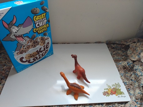 Several dinosaurs checking out a box of Cookie Crisp cereal.